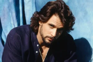 A vintage 1990s portrait of actor Robert Kelker-Kelly with long, thick, flowing dark hair and a trimmed beard. He is wearing a dark blue linen shirt with a silver chain necklace, leaning forward slightly against a crumpled blue fabric backdrop while looking intently at the camera.